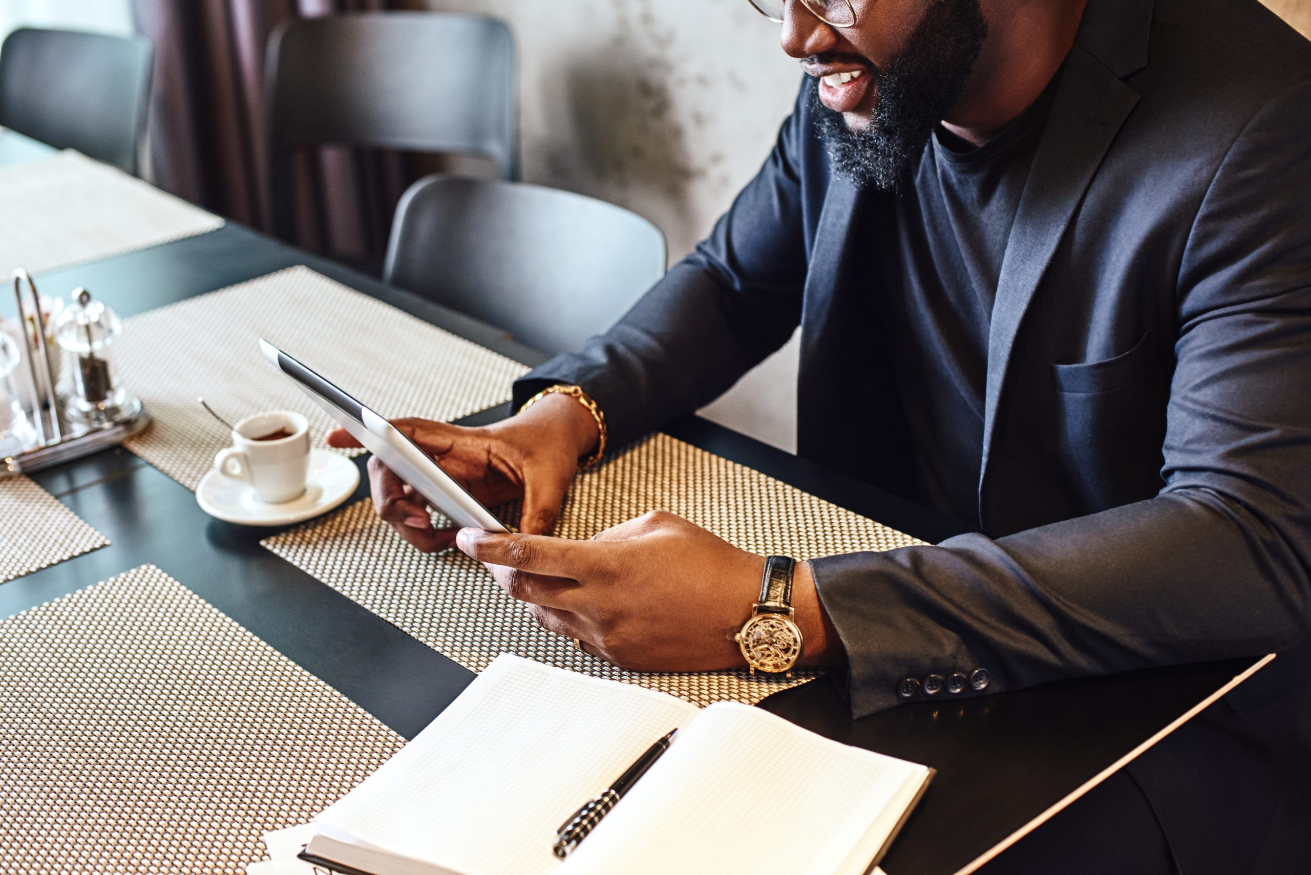 Portrait of focused African-American office manager in dark-blue jacket and glasses, sitting at cafe table with tablet pc, reading documents and smiling. Coffee is on the table. Cozy interior